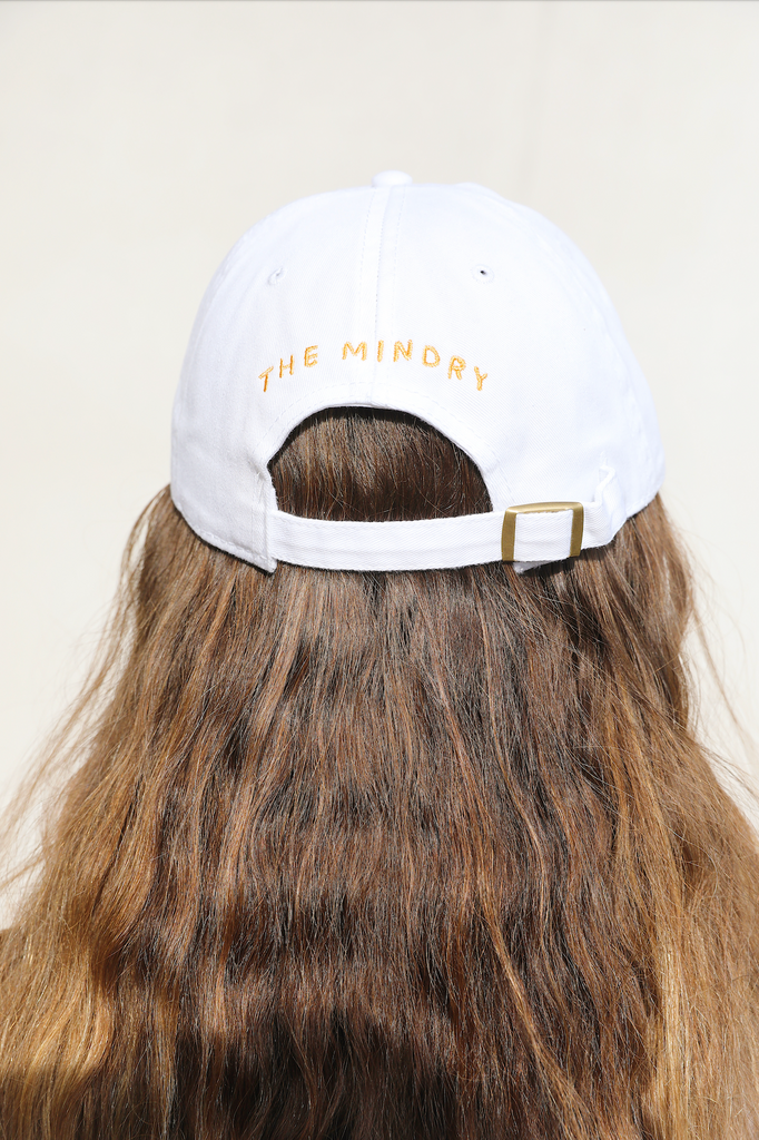White colored dad hat featuring text "The Mindry" on the back. Worn by model facing away from the camera view.