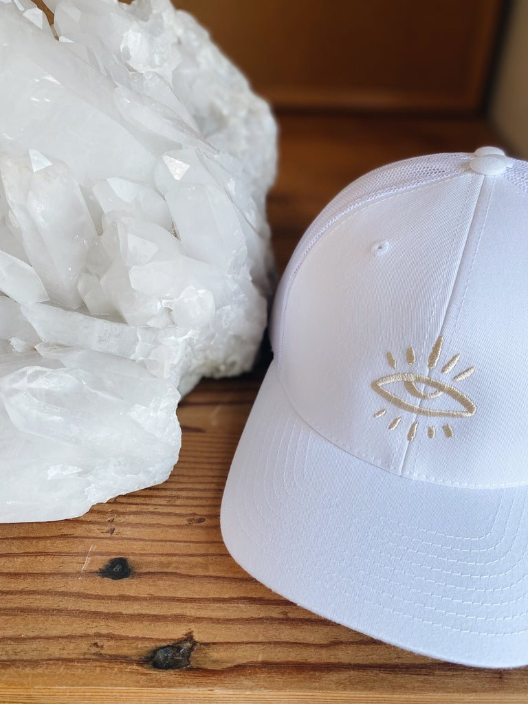 White and sand-colored trucker hat featuring The Mindry logo on the front positioned near a gemstone display.