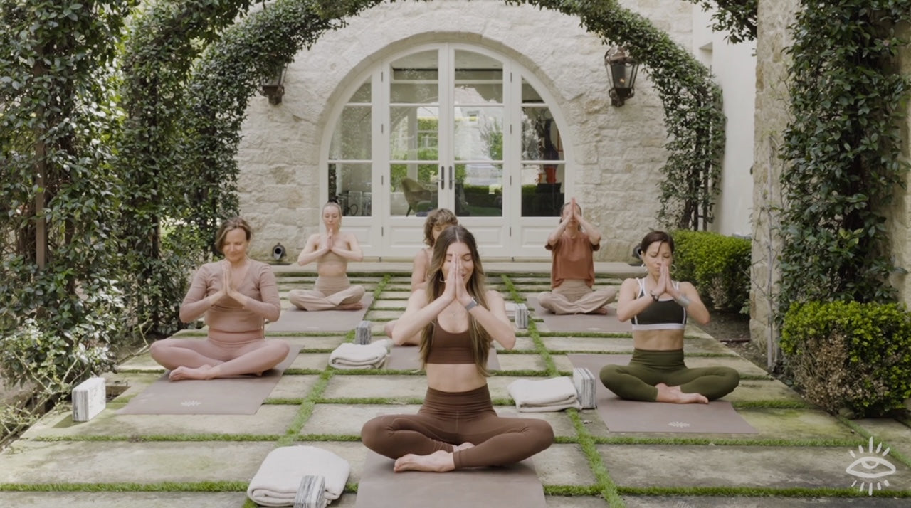 A small class seated in meditation on mats in a green courtyard outside an ivy-covered building