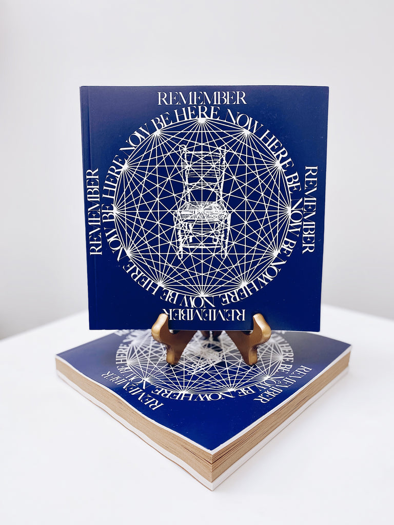 Be Here Now book on display. A blue cover with geometric shapes and a graphic of a chair in the center.
